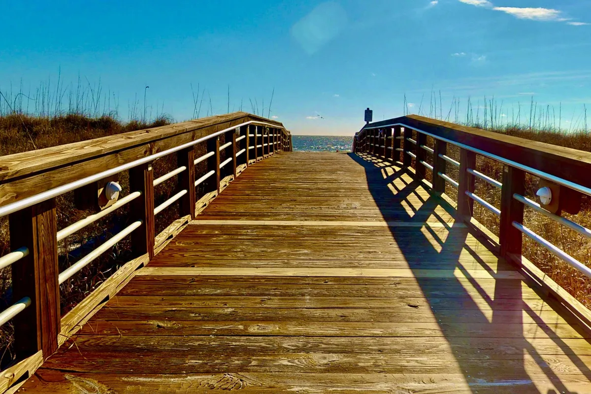 A wooden boardwalk stretches toward the ocean with distant pilings and a serene, open sky, creating an inviting path for leisurely walks. The area is quiet, perfect for couples seeking a tranquil seaside stroll at sunset or sunrise.
