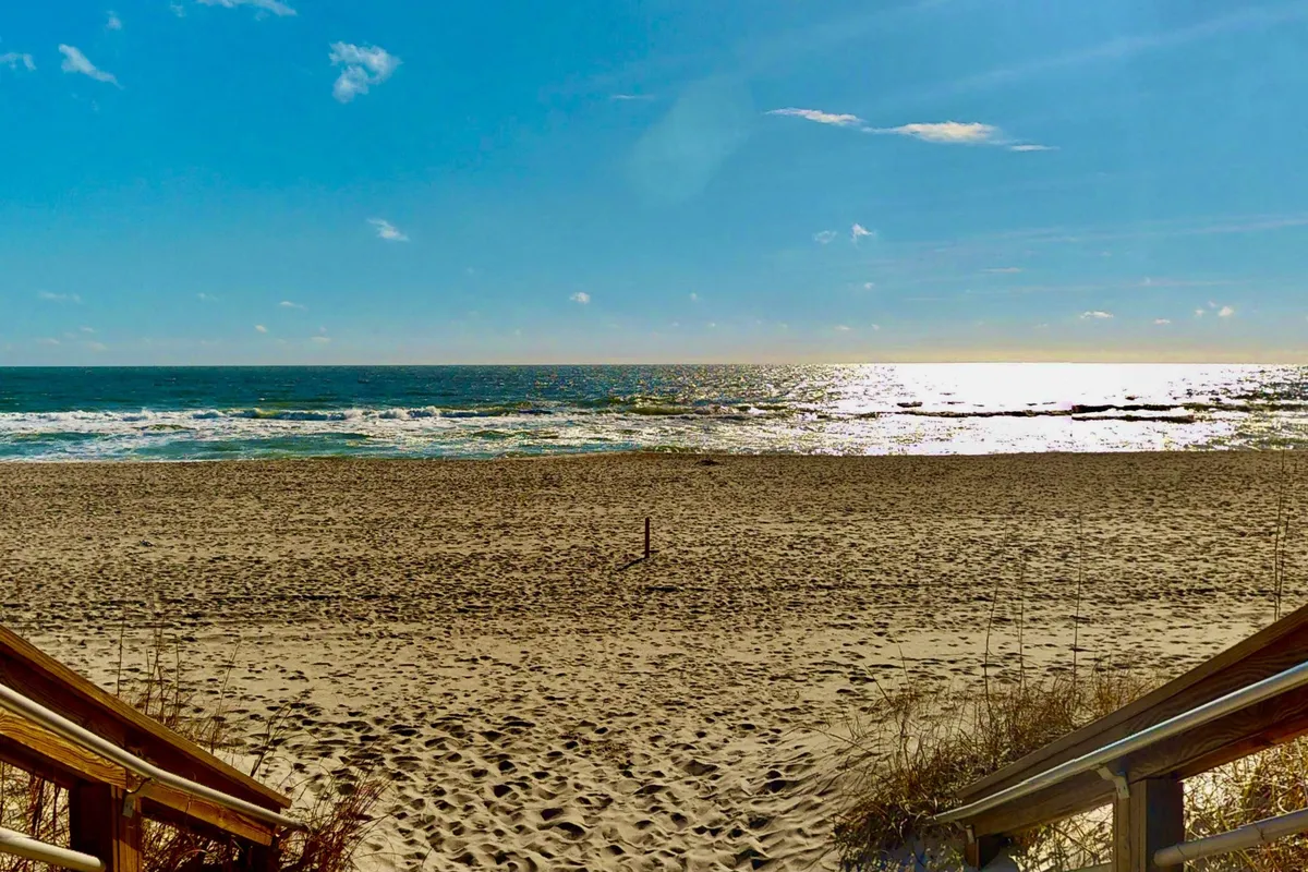 A gently sloping sandy beach meets rolling ocean waves, with wooden steps leading directly down to the shore. The scene is bright and inviting, ideal for families to build sandcastles, swim, or stroll along the shore. Children and adults alike can easily imagine themselves enjoying classic beach activities here.