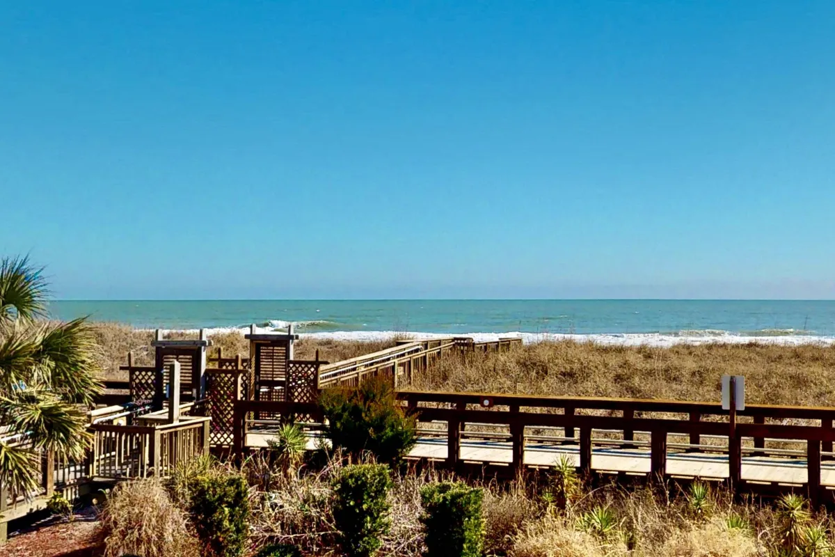 A wide wooden boardwalk bordered by seagrass and palm trees leads visitors toward the ocean, with soft morning sunlight casting a warm glow. The tranquil scene suggests early walks, collecting seashells, and the gentle start of a beach day.