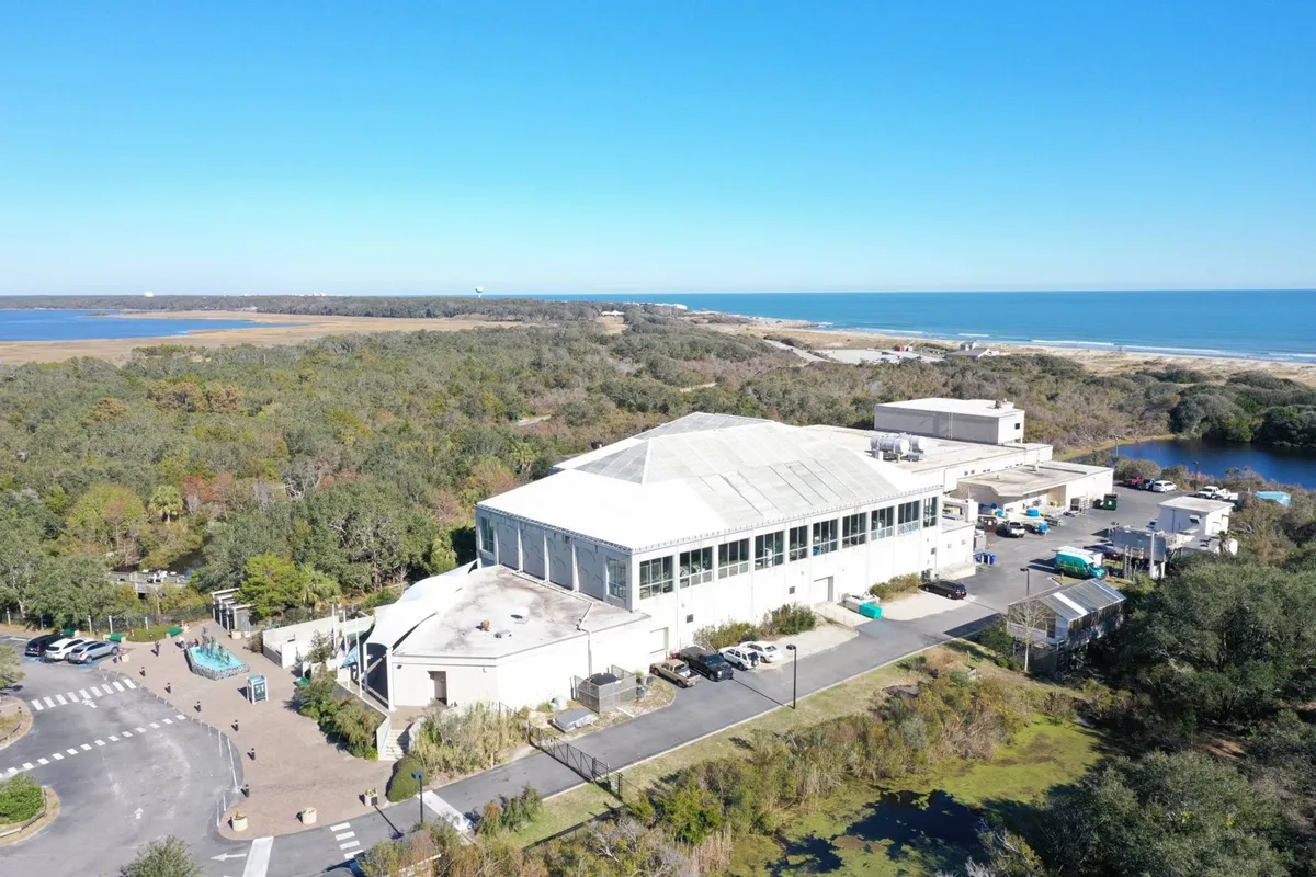 This exterior image shows a large building set among lush greenery with a coastal view in the background. The inviting grounds are landscaped, and the building stands as a focal point, suggesting a community hub or attraction amid a peaceful, natural setting.