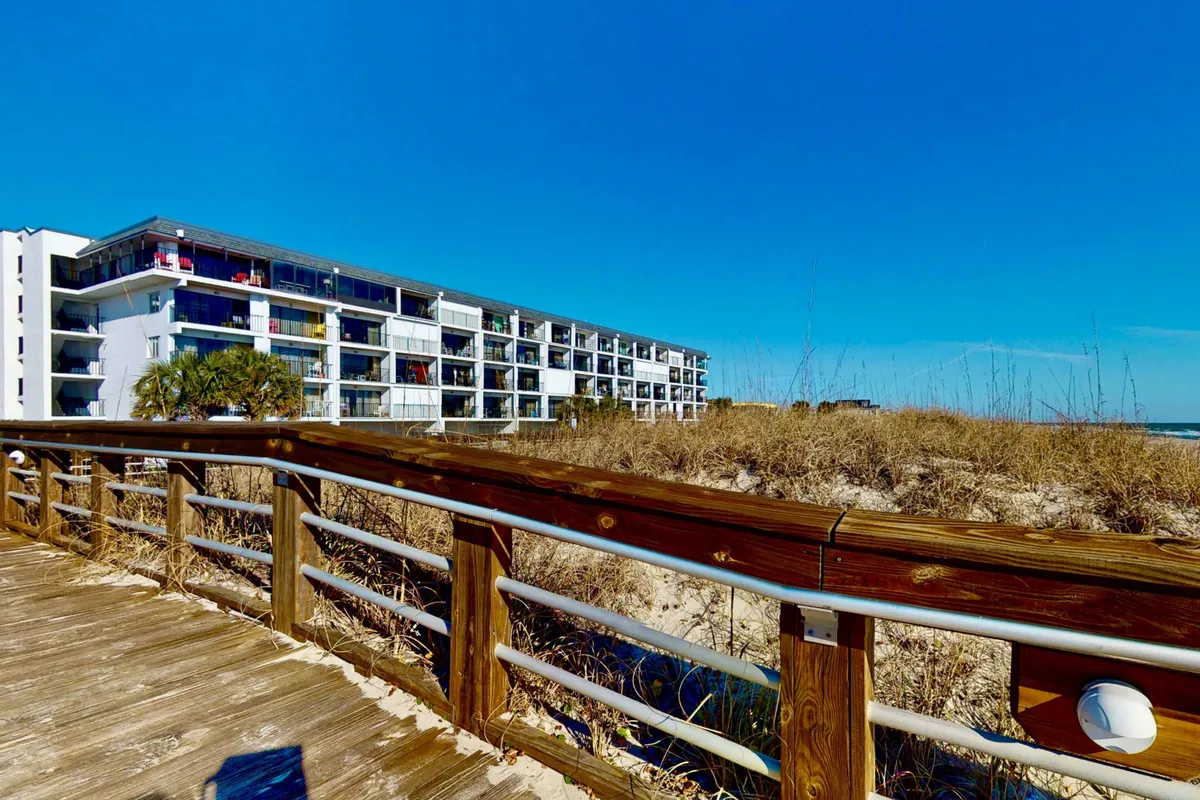 A view of the classic beachfront property with the boardwalk in the foreground and a clear blue sky overhead. People are seen walking along the boardwalk and enjoying views of the sand and ocean, capturing the vibrant coastal atmosphere.