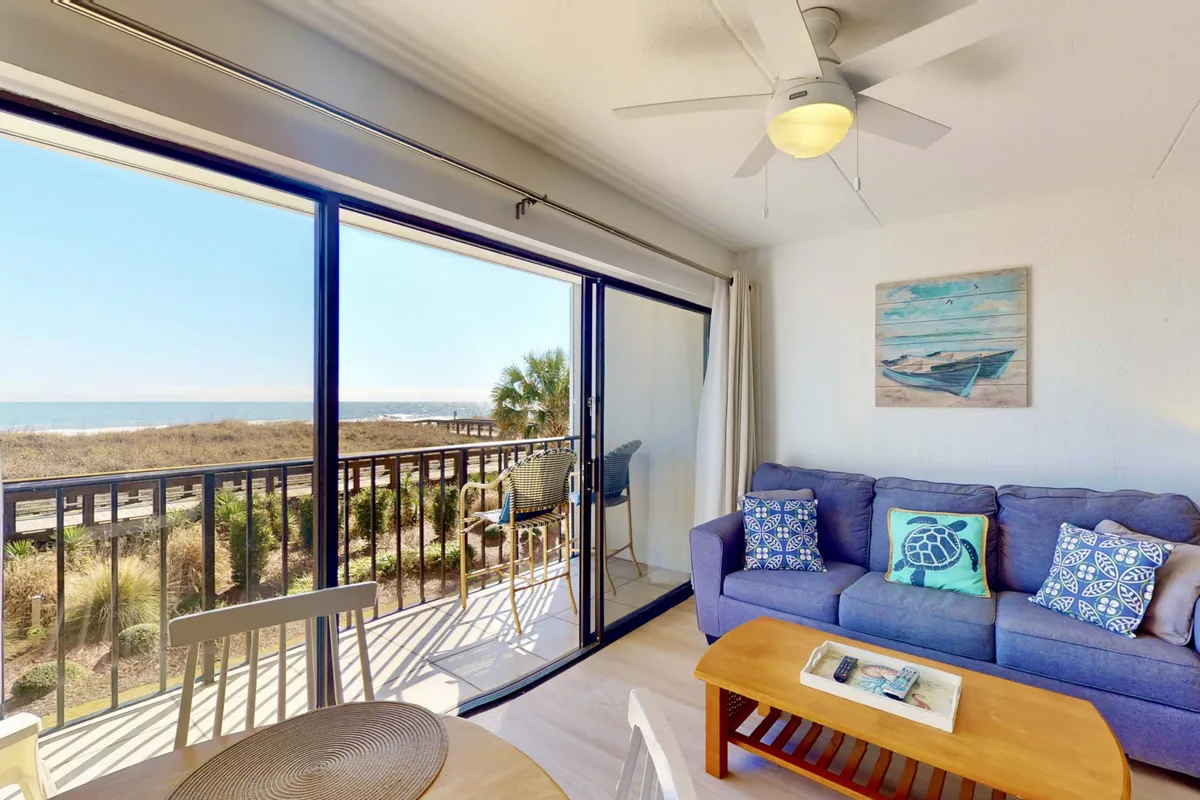A bright, open-concept living space with a comfortable sofa adorned with colorful cushions, a wooden coffee table, and wall art. Large windows frame stunning ocean views, filling the room with daylight and a sense of openness.