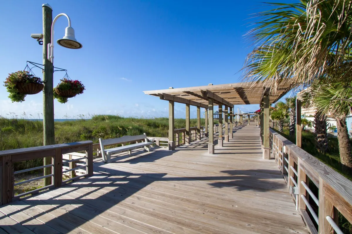 A cheerful boardwalk scene with wooden decking, vibrant hanging flower baskets, and sunny skies. Palm trees add a tropical flair, and the open, social atmosphere invites exploration. Families can easily imagine strolling, enjoying treats, or capturing memorable photos here—mirroring the lively spirit of the nearby Carolina Beach Boardwalk and amusements.