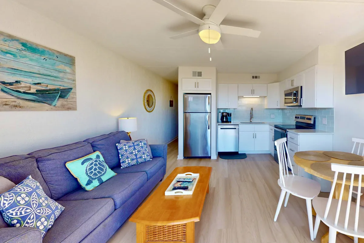 A bright and inviting open-concept interior featuring a cozy sofa with decorative pillows, a dining table set for four, and a fully equipped kitchen area in the background. Natural light illuminates the space, offering glimpses of the ocean through large windows.