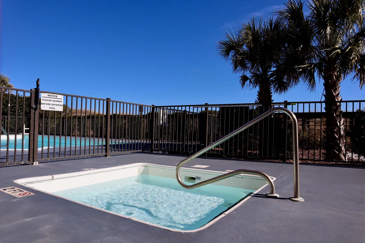 A sparkling community swimming pool surrounded by palm trees and a secure fence, with a relaxing spa tub off to the side. The area is sunlit and tranquil, suggesting a welcoming environment for families to swim, play, or unwind together. The pool’s resort-style ambience matches the description of Casa Al Mare’s family-oriented amenities.