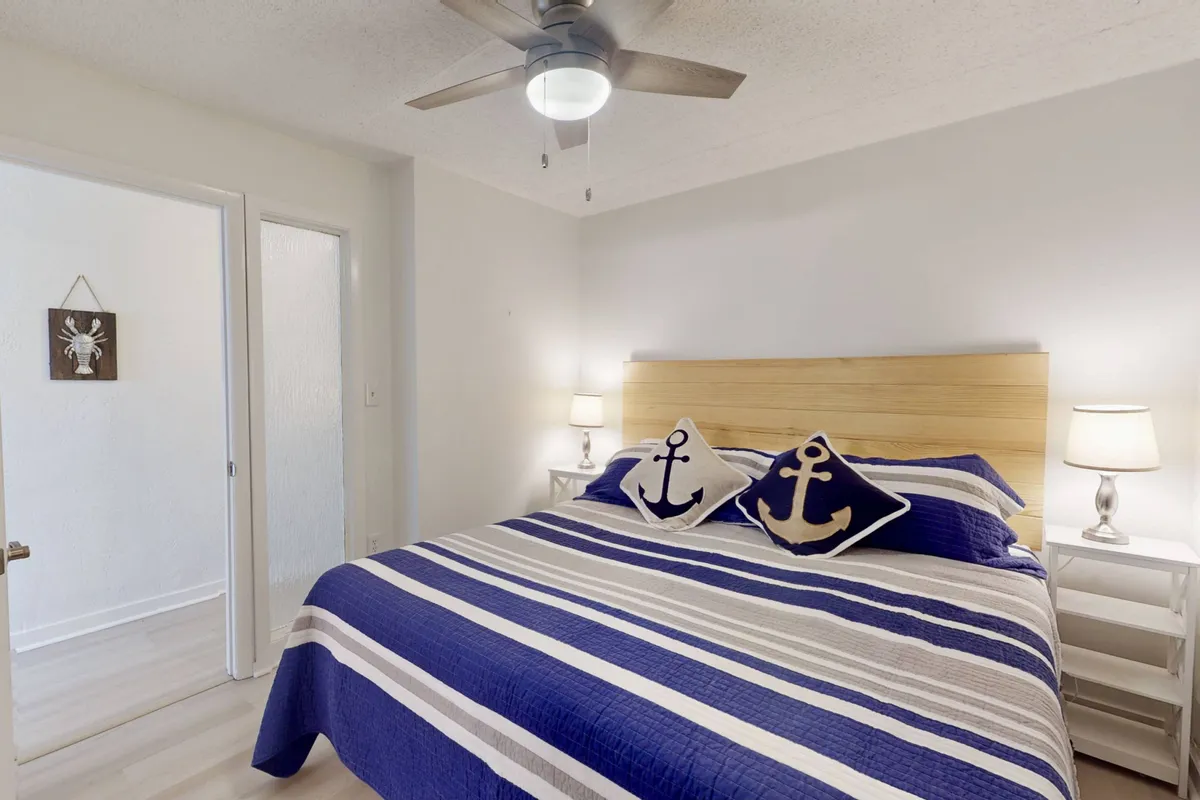 A cozy bedroom with a striped bedspread, anchor-themed pillows, and nightstands topped with soft-glowing lamps. Sunlight filters in, suggesting a bright and peaceful start to the day. The room radiates comfort and tranquility, ideal for restful nights and gentle mornings.