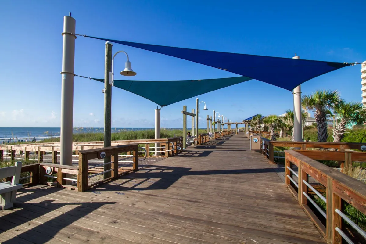 An exterior shot of a wooden boardwalk lined with colorful shade sails, palm trees, and a clear view of the beach and ocean beyond. The scene is tranquil, inviting strolls or biking, with plenty of seating and spaces to pause and enjoy the view.