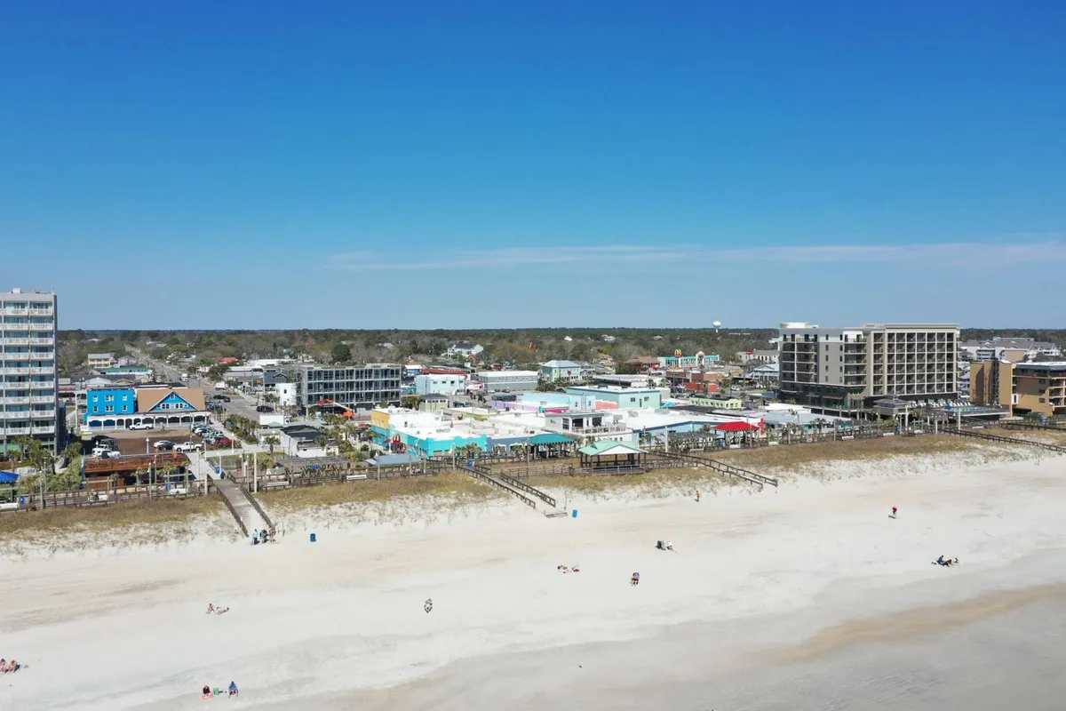 A lively beachfront scene featuring colorful buildings, the bustling boardwalk, and people enjoying the beach and nearby shops and restaurants. The atmosphere is vibrant and full of energy, reflecting the fun and accessibility of the Carolina Beach Boardwalk experience.