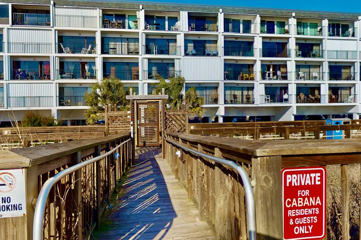 This image shows a wooden walkway lined with palm trees leading from the condo building's balconies directly toward the beach. The balconies are furnished with chairs, and the entire scene feels tranquil and welcoming, emphasizing easy access from living spaces to the boardwalk and beach.