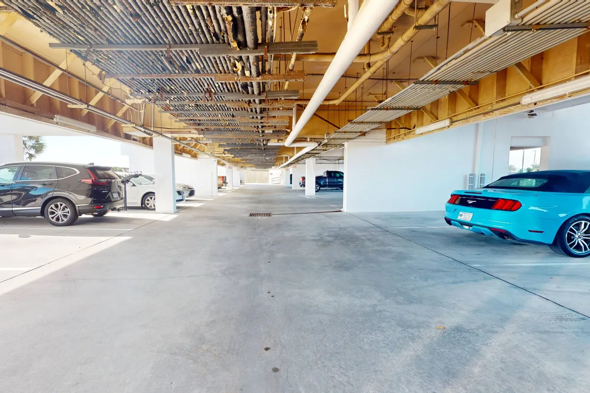A practical covered parking garage with several cars parked, exposed ceiling pipes, and concrete flooring, demonstrating the convenience and safety of onsite, sheltered parking for residents and guests.