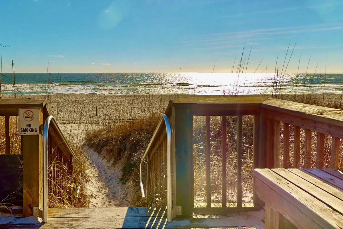 Wooden steps descend through dune grass directly onto the sandy beach, with gentle ocean waves visible beyond. The setting suggests the start of a secluded walk or the perfect spot for intimate beachside moments.