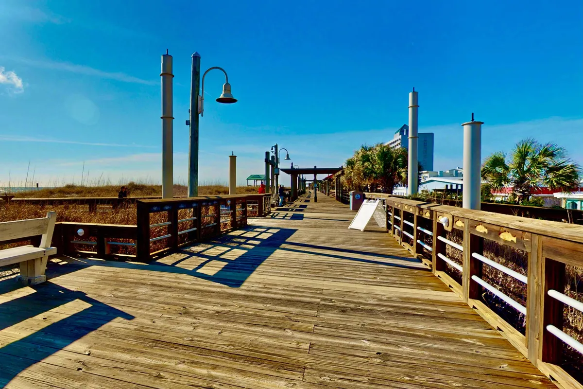 A welcoming exterior shot of a wooden boardwalk lined with lampposts, surrounded by coastal greenery and leading towards the beach. Beach access signs and benches invite visitors to linger and soak up the relaxed seaside vibe.