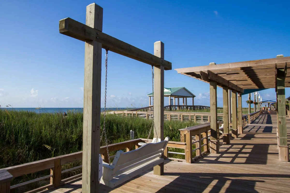 A swing set on a pathway overlooks the ocean, surrounded by pavilions and lush landscaping. The inviting swing offers a perfect spot to relax, watch the sunrise, or simply enjoy the ocean view and boardwalk ambiance.